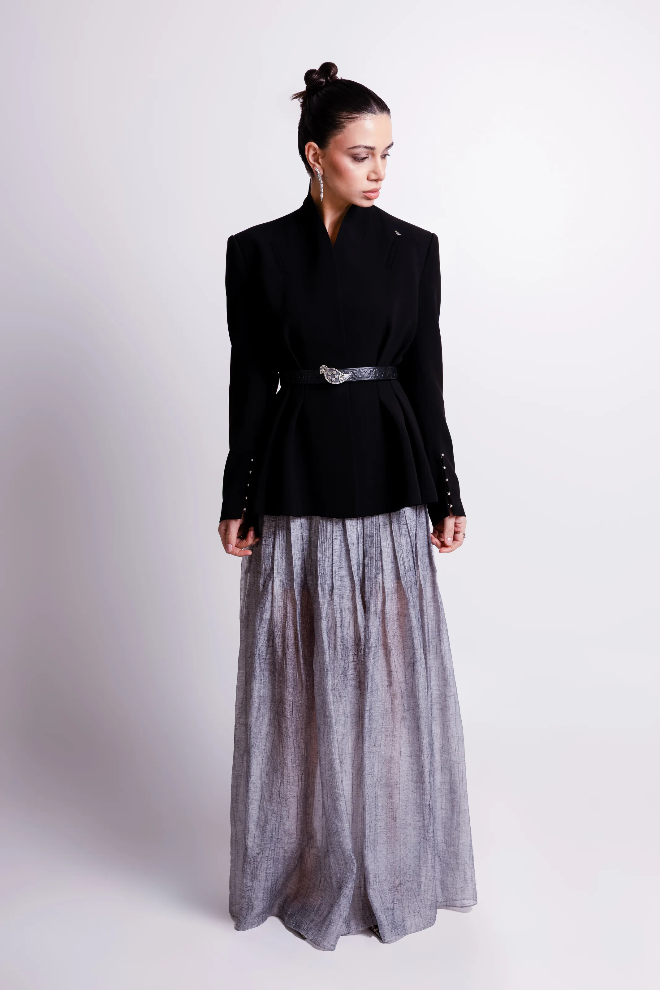 Black jacket with a standing collar and flared hem, belted with a silver bird buckle, over a long sheer grey skirt