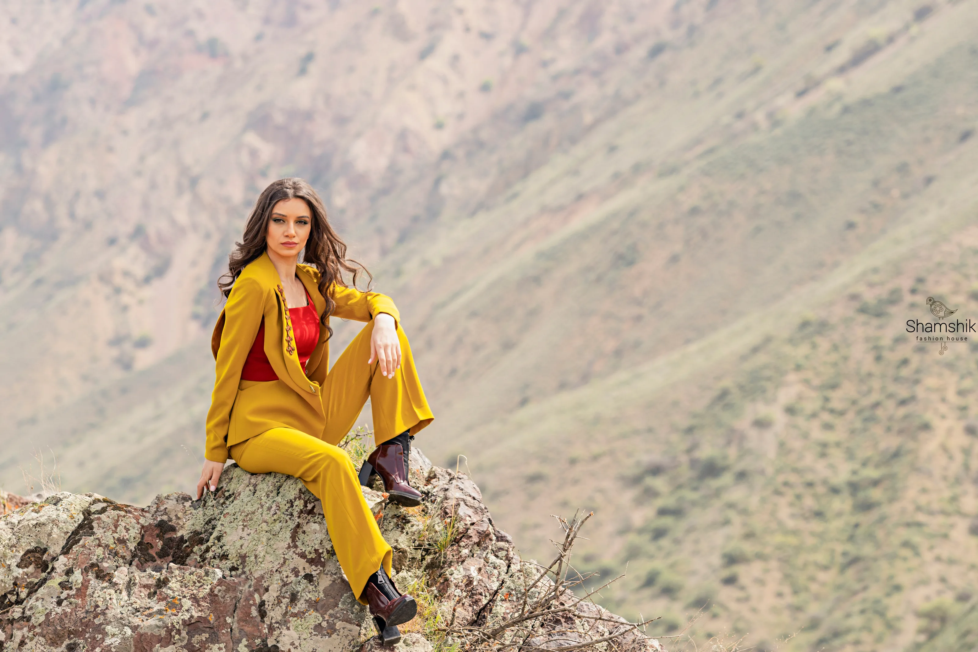 The mustard suit seen full length, seated on rock against a mountain slope