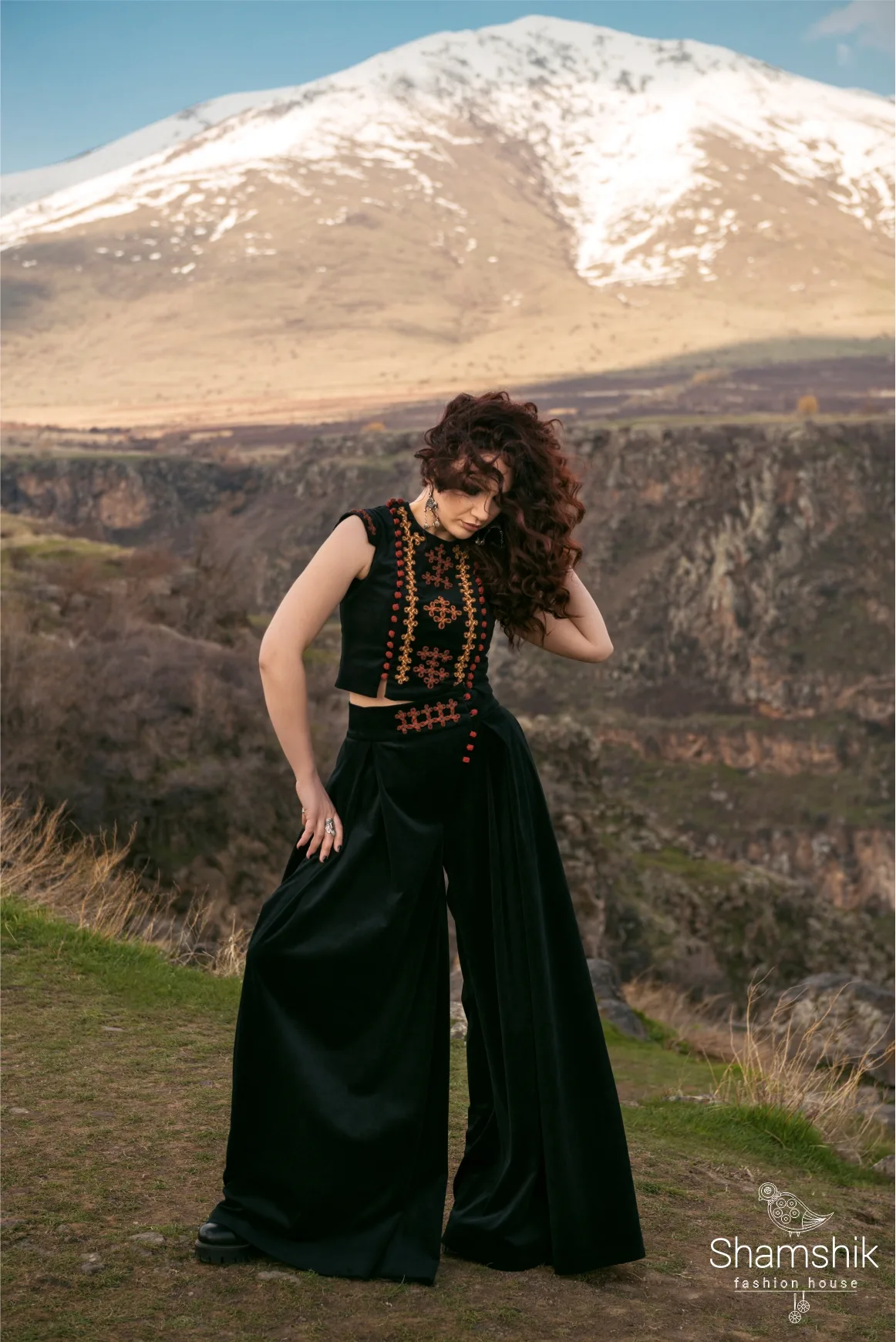 Black sleeveless top and wide palazzo trousers with amber and red cord embroidery, before a snow-covered mountain