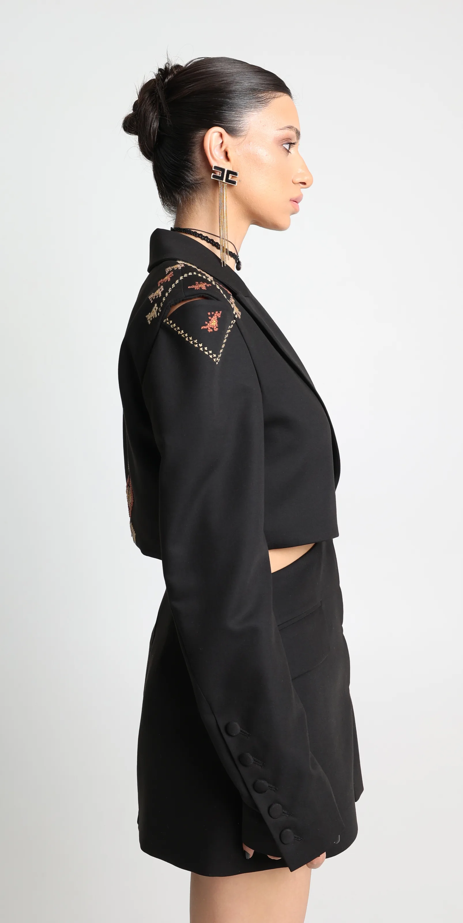 Cropped black tailored jacket with peak lapels, hand cross-stitch at the shoulder in rust, gold and cream, worn with a black mini skirt