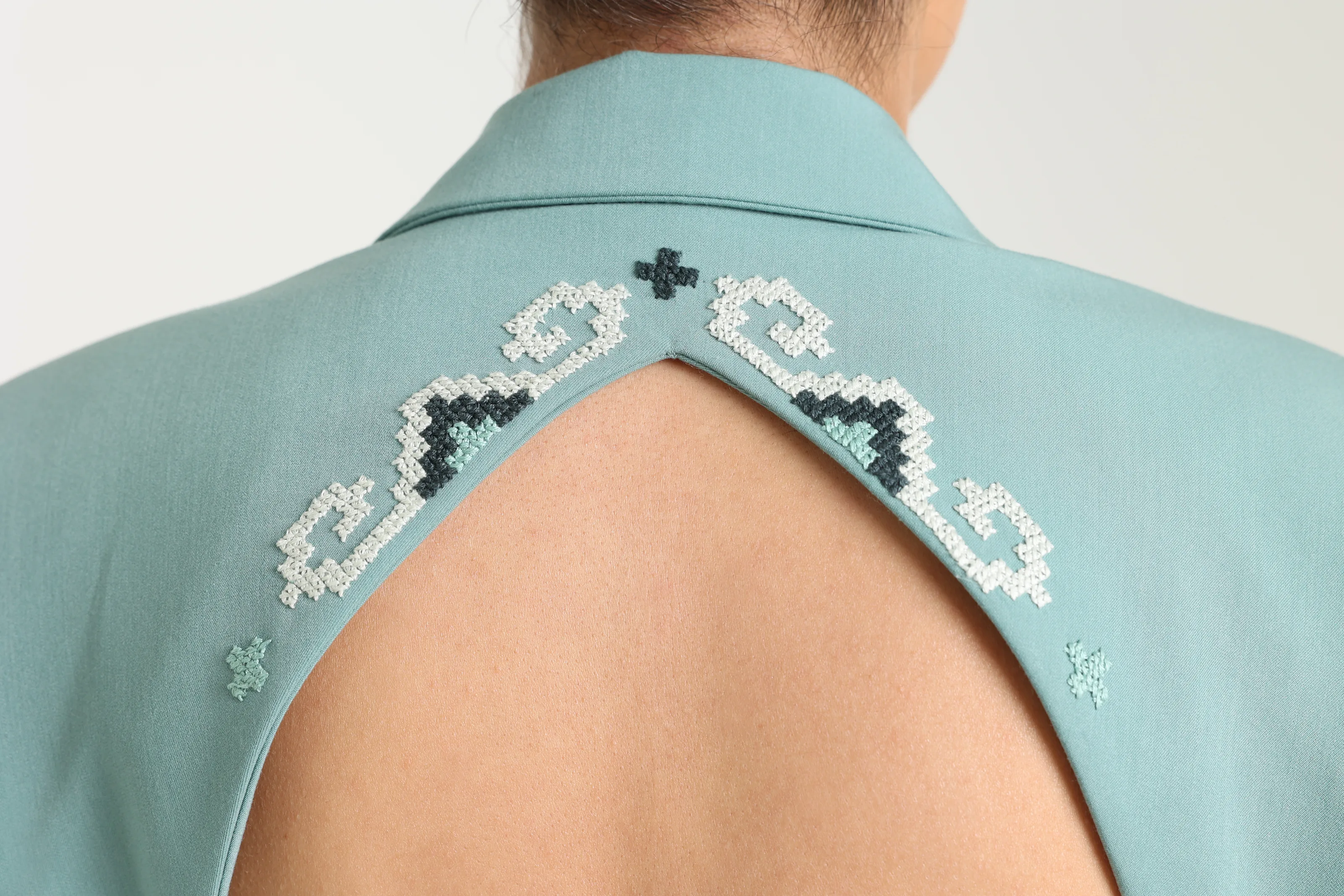 Close view of the open back of the sage jacket, collar and opening edged in white, dark teal and mint cross-stitch